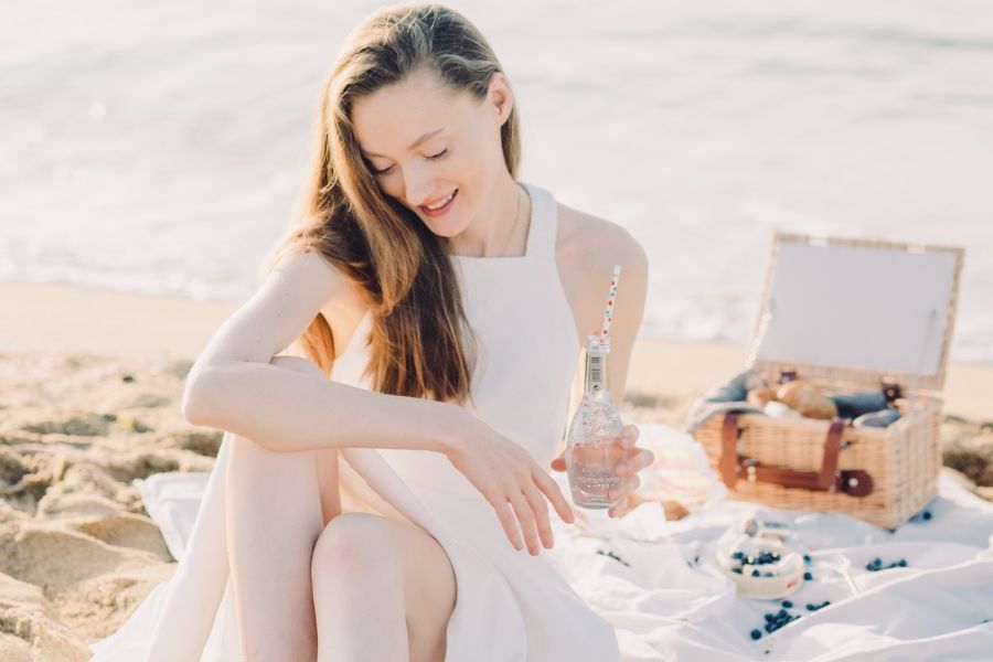 Girl with a water bottle and picnic basket, sitting on the beach in white dress, practicing one of the best solo date ideas. 