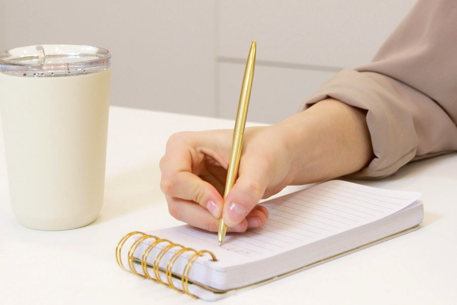 a hand on white table, writing positive affirmations for mental strength in a notepad with a golden pen