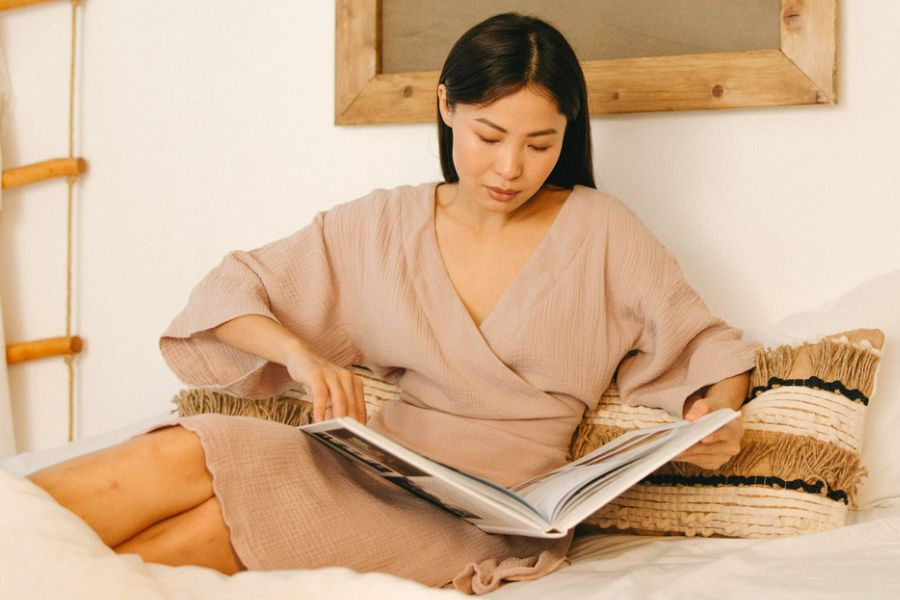 woman in beige dress sitting in bed, reading a book as part of her self-care night routine 