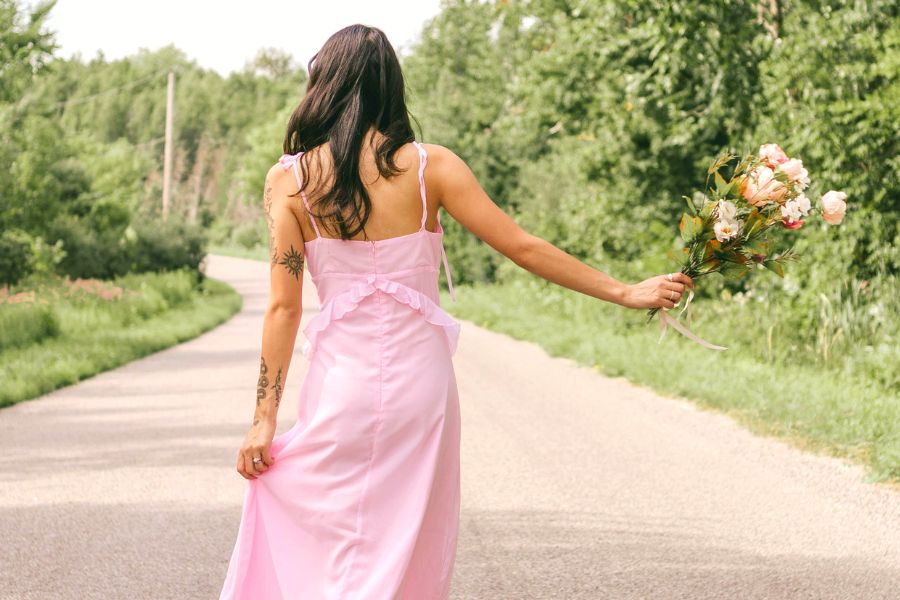 woman in a pink dress, holding  a bouquet, walking on road with her back to us, indulging to mindful habits to improve her mental health 