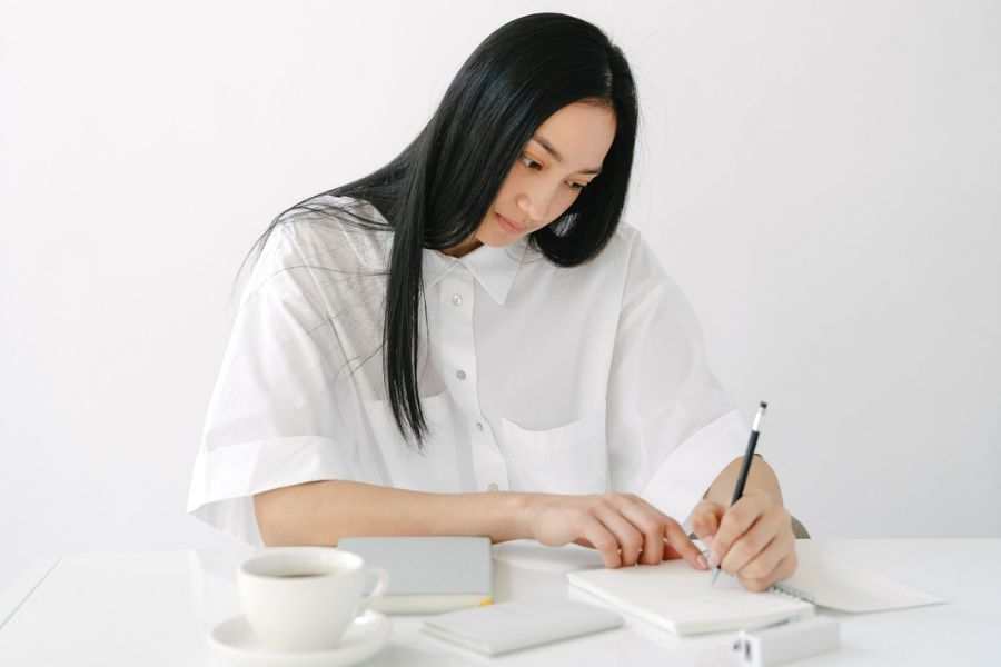 woman in white shirt writing in a notepad, with stapler and coffee on table, indulging in good Monday habits 
