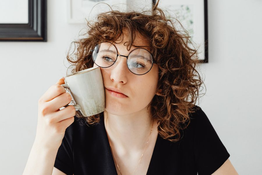 woman holding a mug to her cheek, wearing tilted glasses, thinking how to stop procrastinating