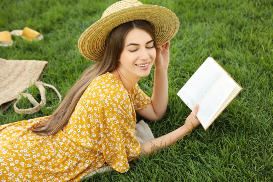 a happy woman wearing yellow sundress and hat, lying on grass while reading a book