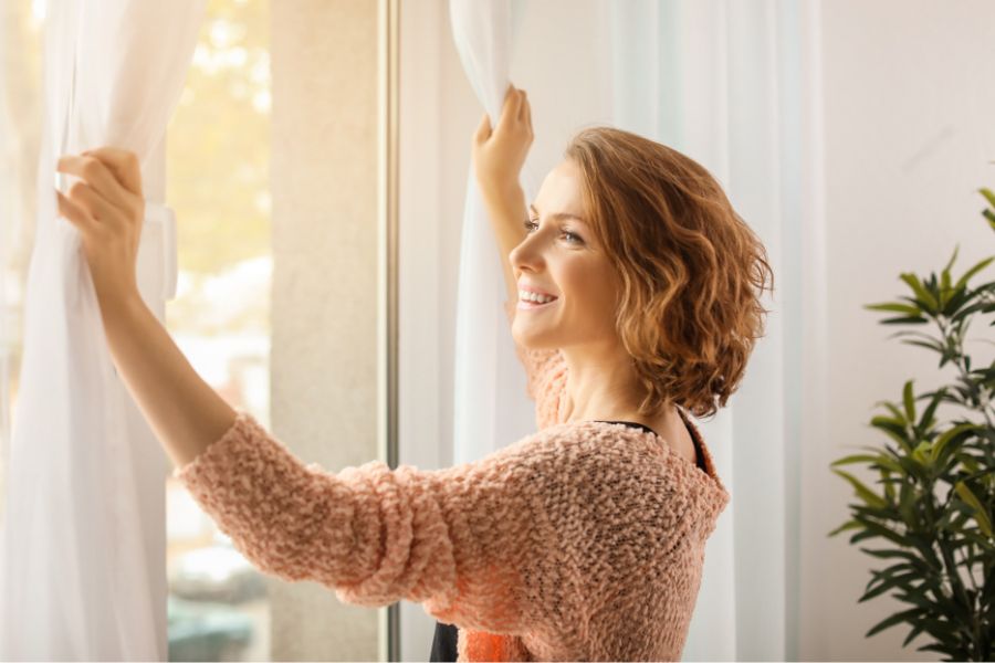 happy woman wearing a peach sweater opening curtains on a fall morning