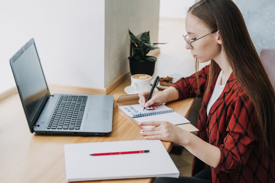 woman writing her goals in a spiral notebook, laptop open in front of her, coffee on table