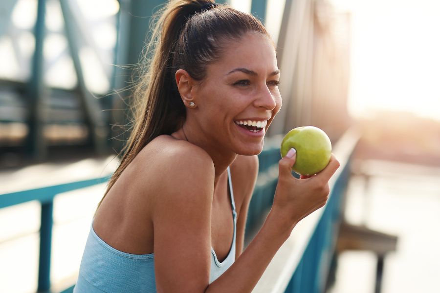 laughing woman eating green apple in workout clothes standing on a bridge