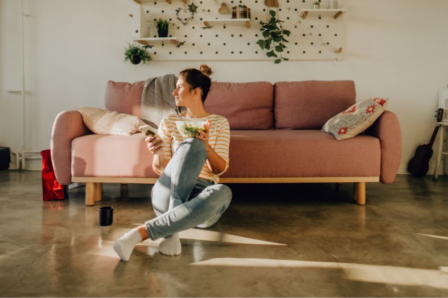 woman sitting on floor next to sofa with remote in hand while eating salad looking outside