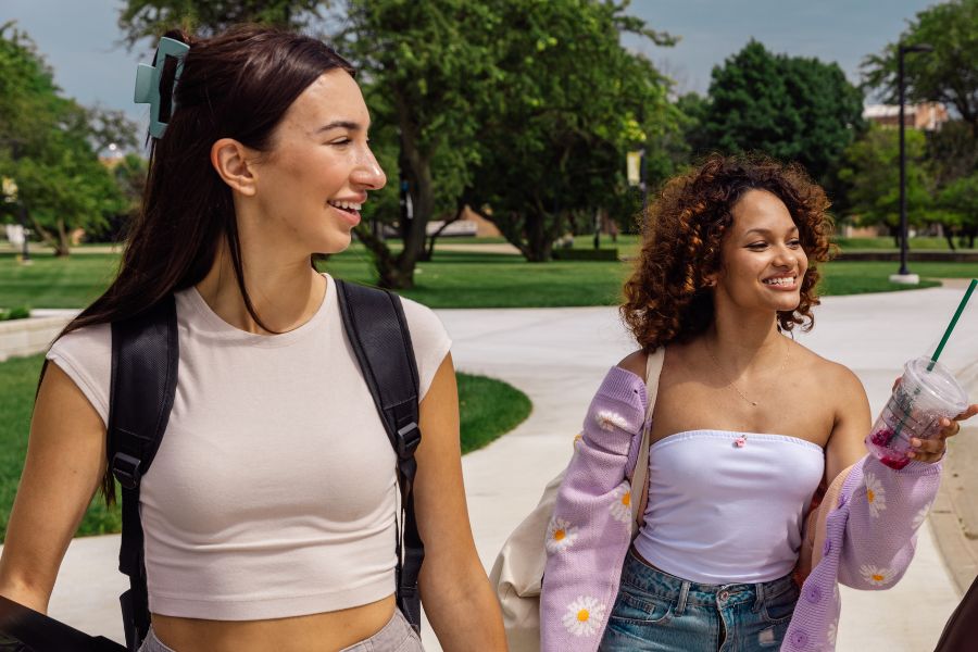 two smiling women walking together, one with a backpack, one with a drink in hand
