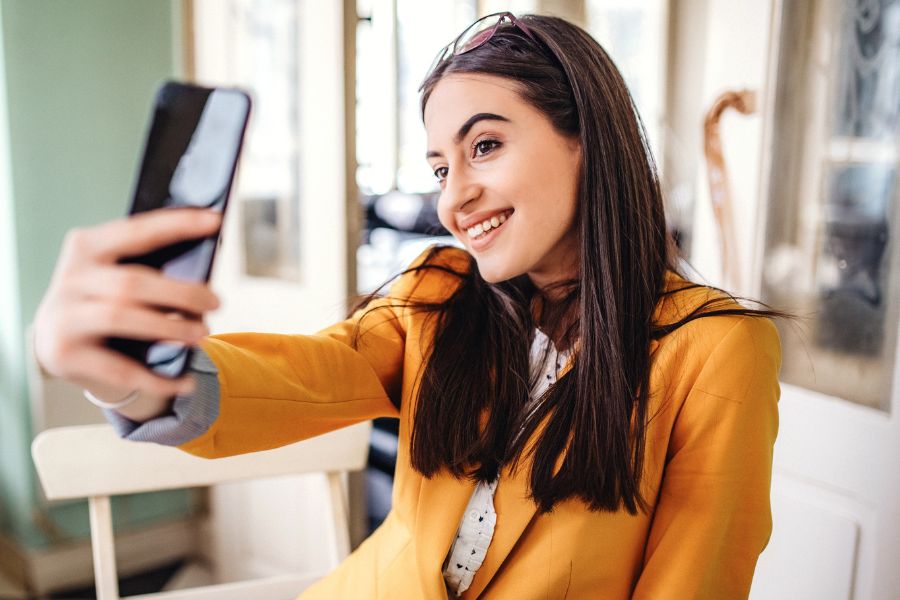 woman in yellow blazer with glasses on head clicking selfie while smiling huge