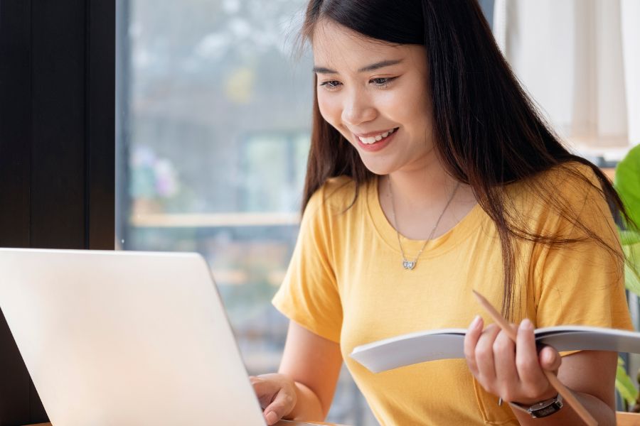 woman in yellow t-shirt smilng while working on laptop, holding notebook and pencil in hand