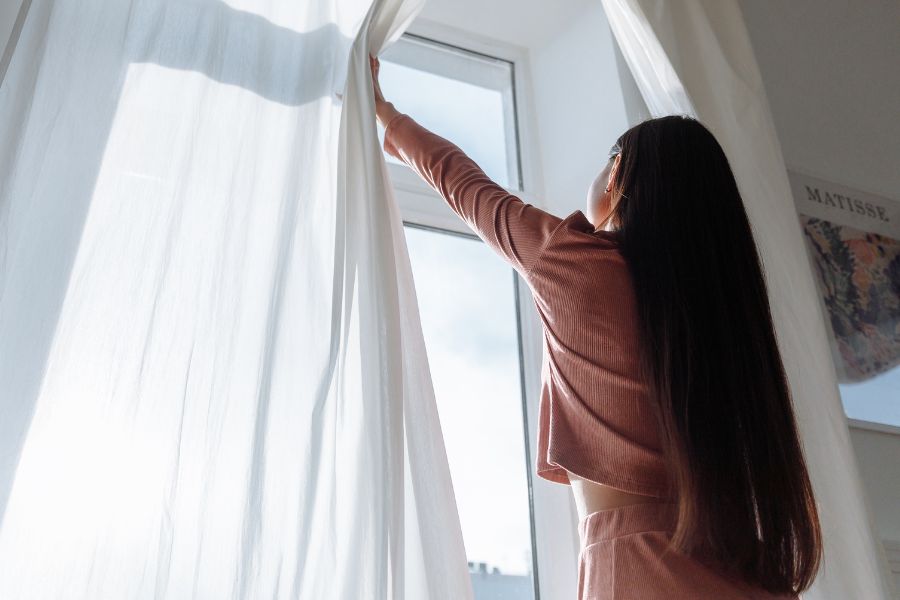 back view of woman opening curtains and letting sunlight in