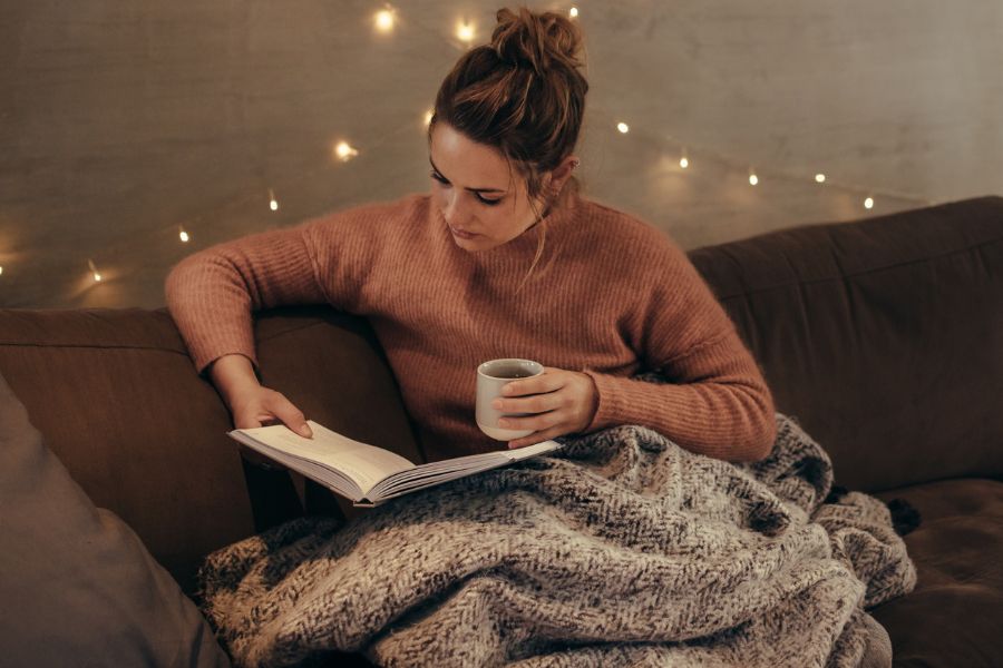 woman reading with tea in hand and blanket on legs, fairy lights behind