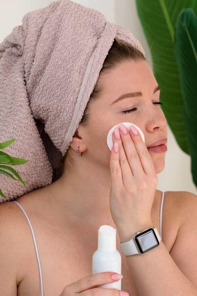 woman with towel on head, swiping cotton pad on face with eyes closed, holding a skincare bottle in hand