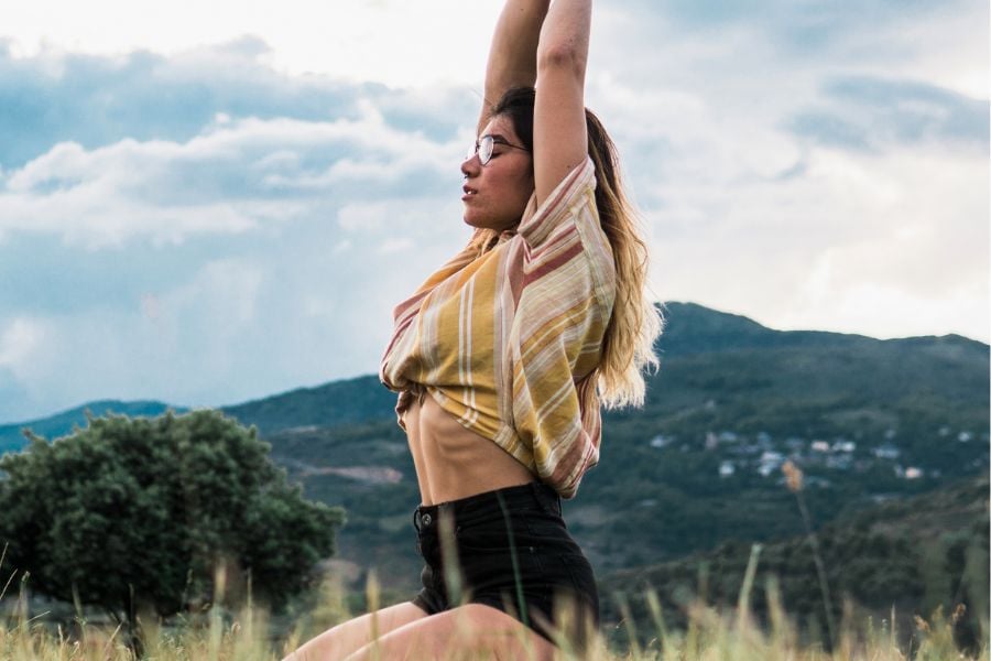 woman stretching her arms up while sitting in grass on her knees, with mountain in background