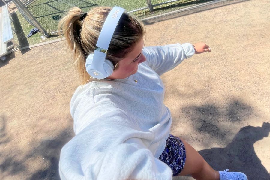 woman looking down while walking in sunlight, wearing white headphones, grey sweatshirt, blue shorts, white sneakers