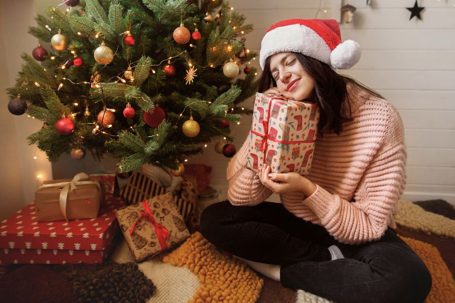woman hugging gift next to a christmas tree, wearing a santa hat