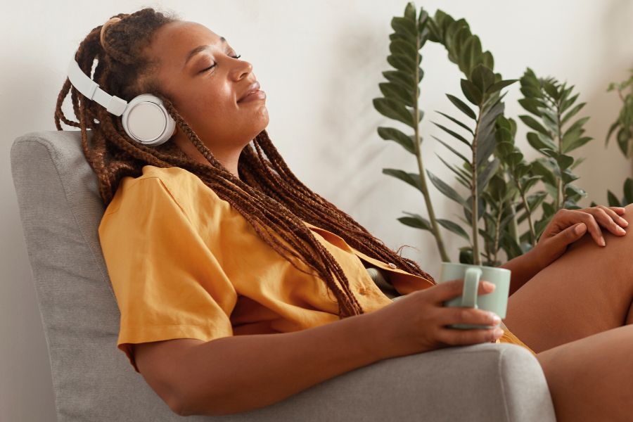 woman in yellow tshirt relaxing on couch with eyes closed, white headphones on, holding cup in hand