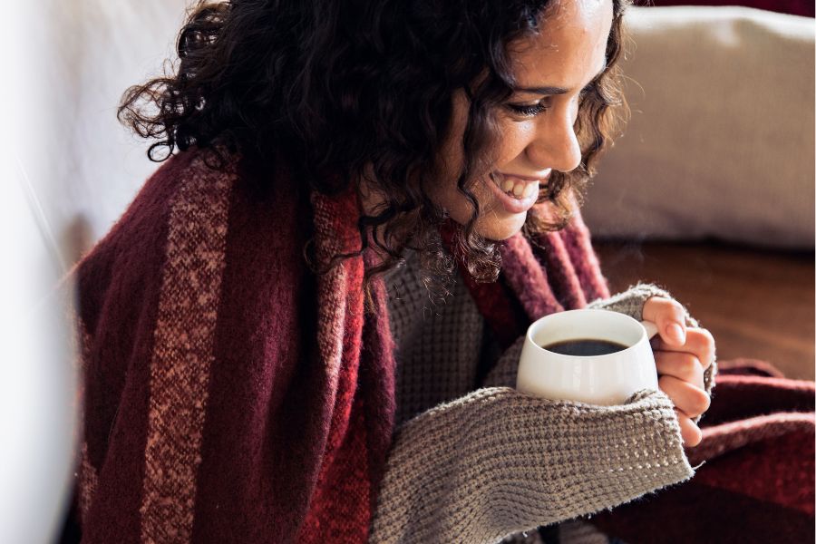 woman with curly hair smiling wide holding a coffee mug, wearing red shawl and sweeater