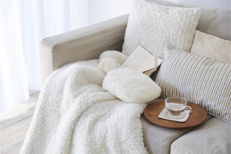 sofa with white blankets, throw pillows and cushions, a book, and water in a transparent cup placed in a wooden round tray
