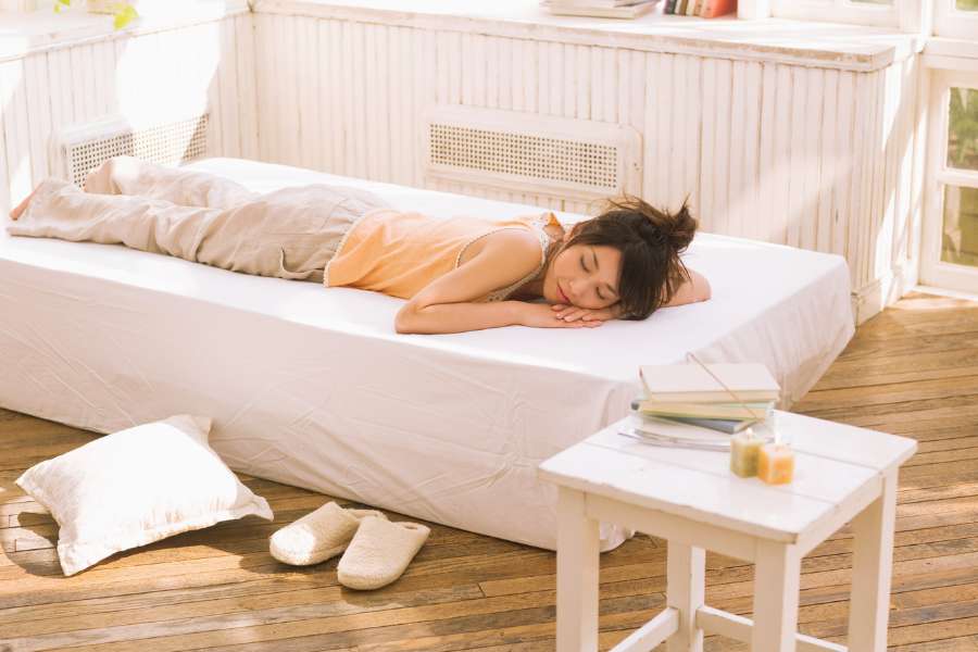 woman lying on her stomach in bed with hands under chin, eyes closed, sunlight entering room, pillow lying on floor, nightstand drawer with candles and journals