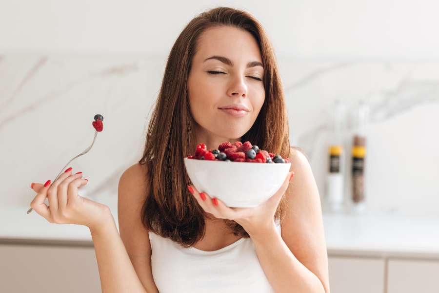 woman holding a big white bowl full of blueberries and strawberries in one hand, and a fork with berries in other hand, eyes closed in joy