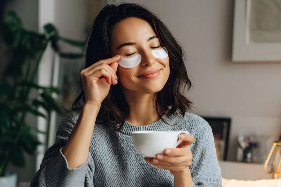 woman with shoulder length hair, wearing gray sweater, smiling while putting on under-eye mask, holding a cup in hand
