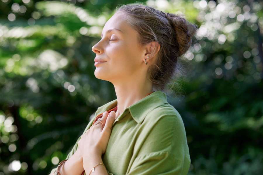 woman in green shirt holding hands over heart, eyes closed, hair in a bun, blurred trees in the background