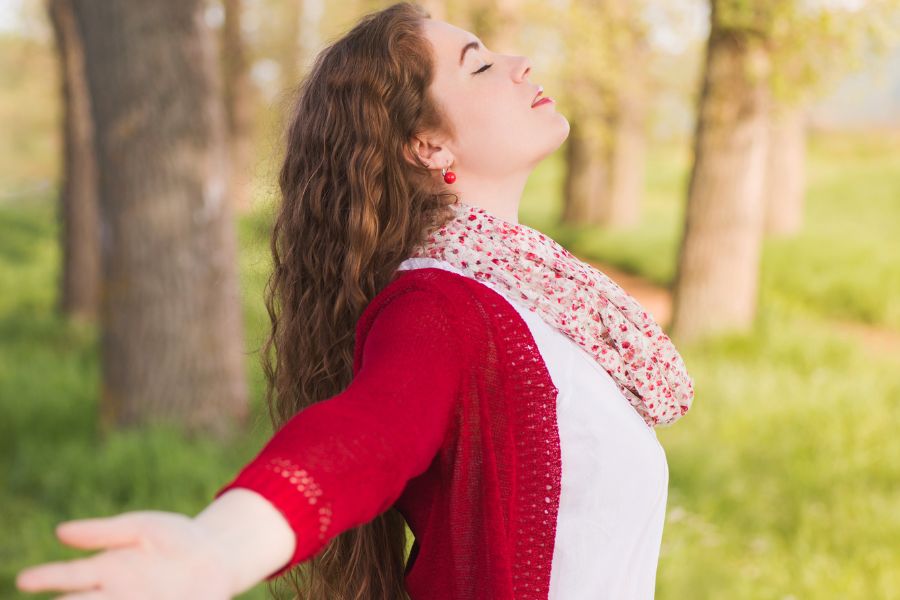 side view of a woman standing outdoors with her arms wide open, eyes closed, and looking up, wearing a red cardigan over a white tee, and scarf on her neck