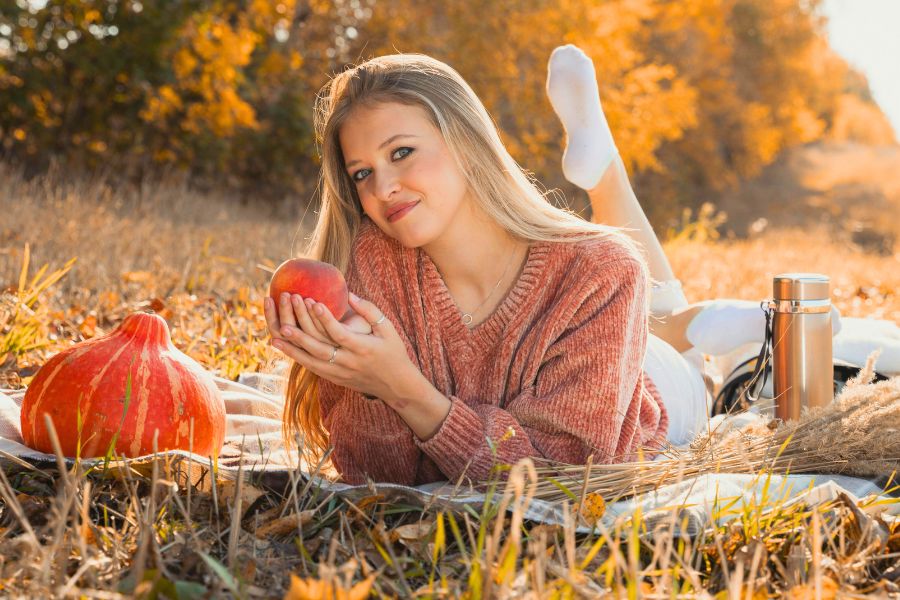 woman on her stomach in a park, on a picnic mat with pumpkin on her left and thermos on her right, holding an apple and smiling, wearing a copper sweater and white socks