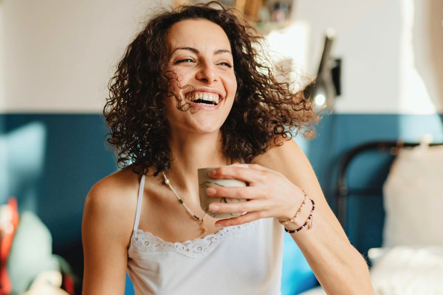 woman with curly hair, wearing a white camisole, laughing freely holding coffee mug in one hand