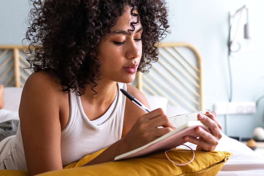 woman with curly hair lying on her stomach in bed, writing in a journal on a yellow pillow
