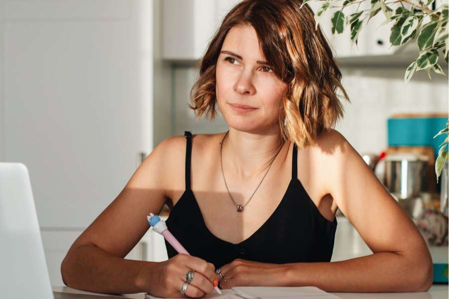 woman with short hair holding pen over notebook, thinking while sunshine falls on her face, wearing a black cami top, a pendant