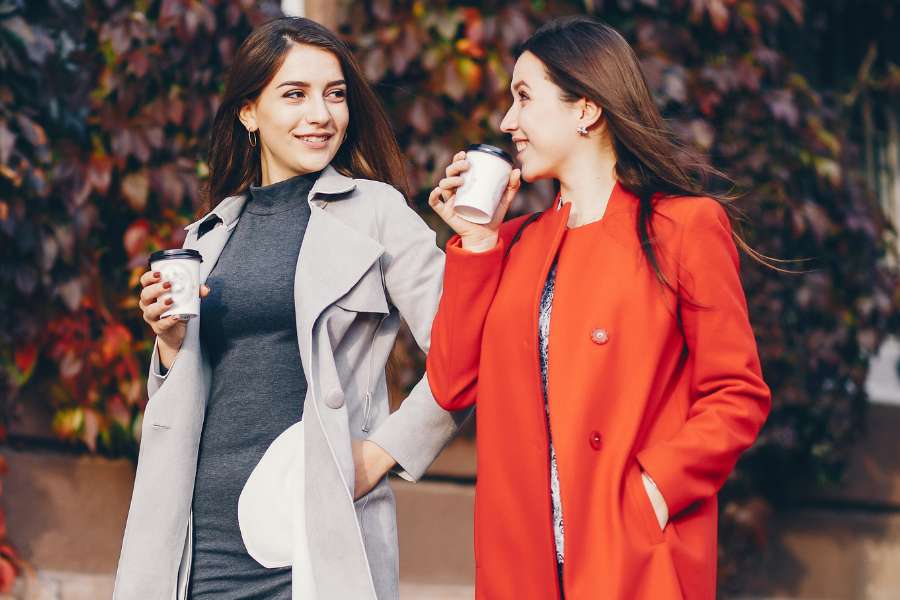 two women friends looking at each other, holding coffee takeaways, wearing overcoats (one grey and one red)