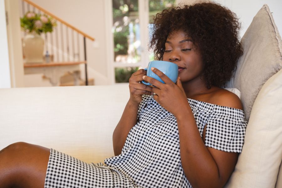 woman on couch sipping from blue mug held in both hands, eyes closed 