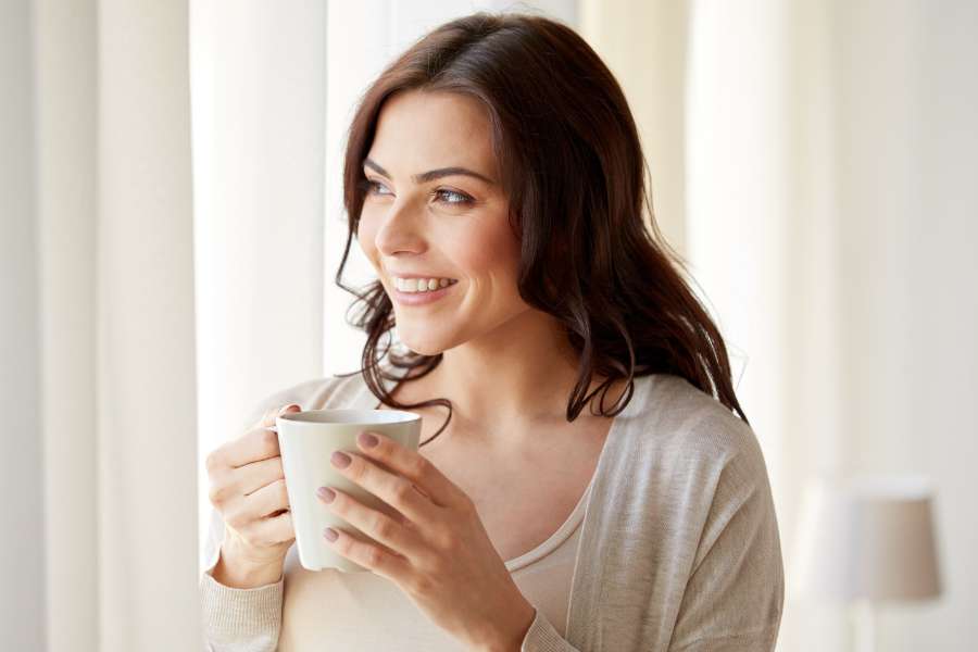 woman smiling looking out the window through curtains, holding a mug in hands