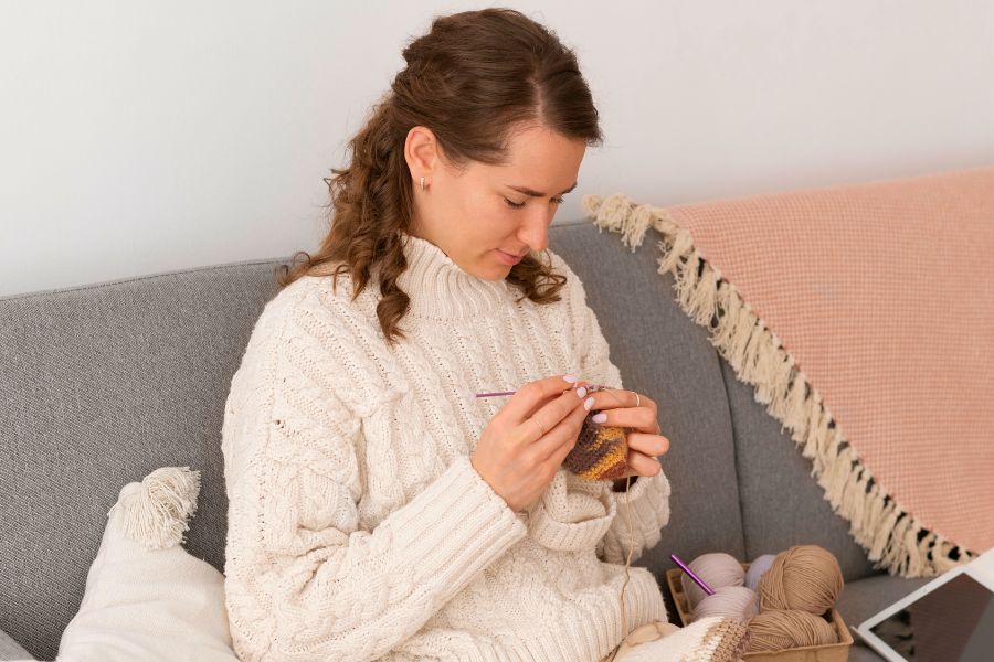 woman sitting on grey couch knitting something brown, wearing cream knit sweater, basket of wool next to her