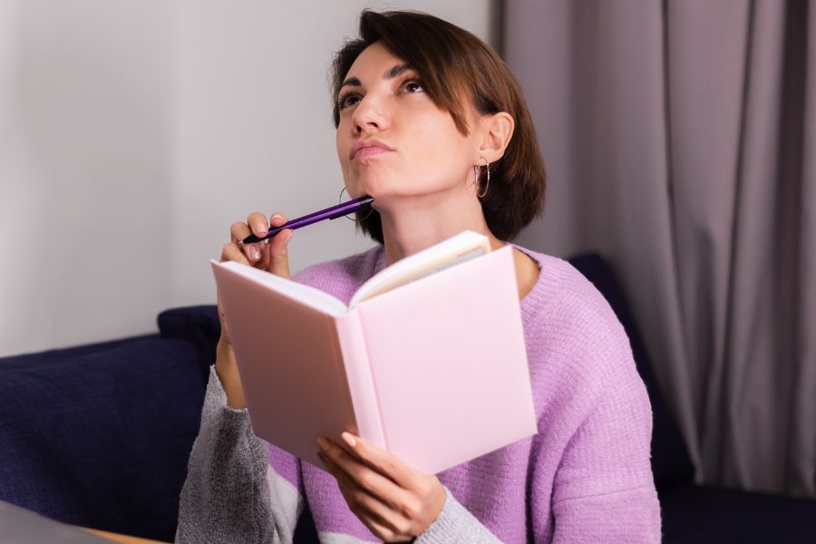 woman wearing lavender sweater holding a diary in one hand, pen on chin in other hand, looking up thinking