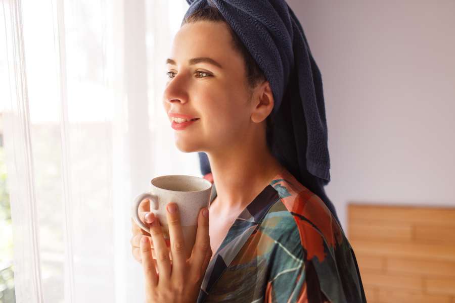 woman with towel on head, holding coffee mug standing near window with white curtain, smiling