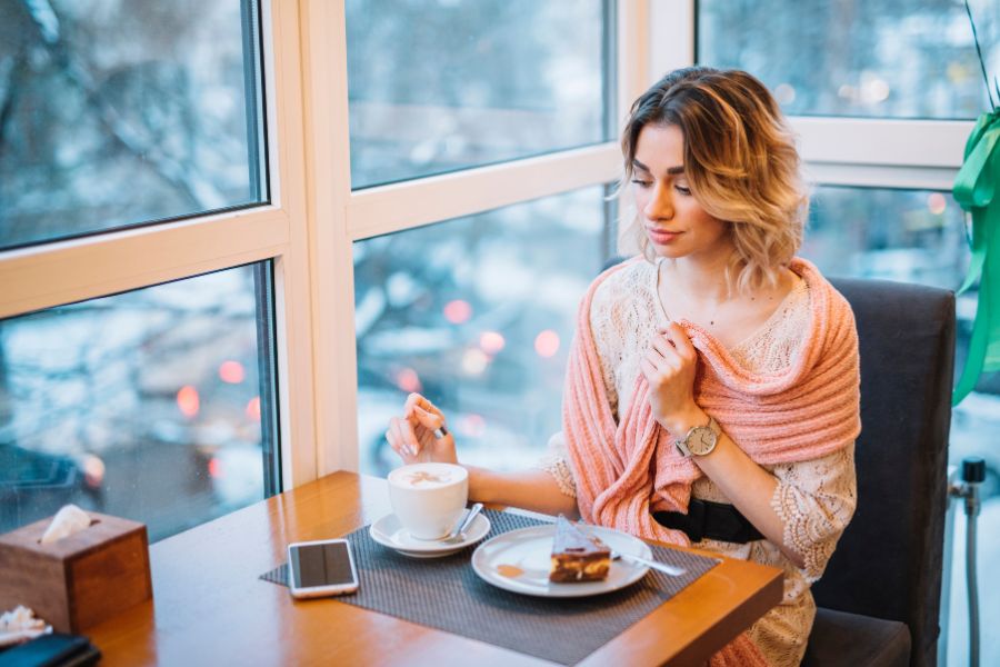 woman sitting in a cafe eating pastry and coffee, wearing a dress, pink acrylic scarf draped on her shoulders