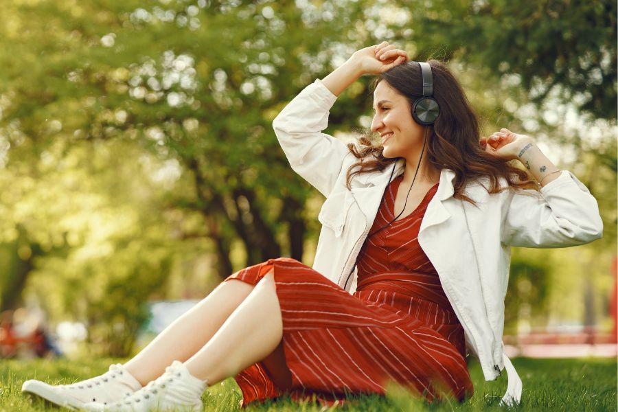 woman sitting on grass with headphones on, hands in air, smiling, wearing orange dress, white jacket and white sneakers