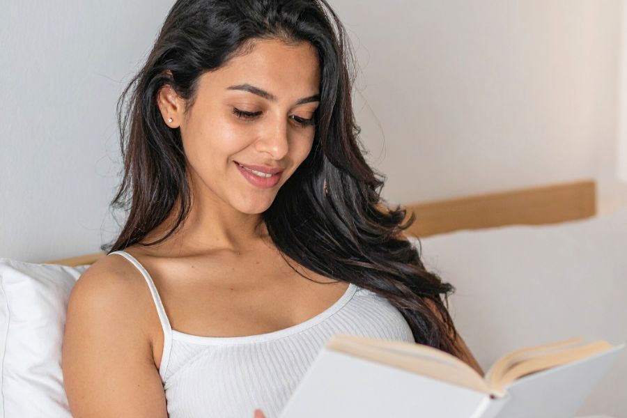 woman reading in bed, hair open, wearing white cami, smiling at book