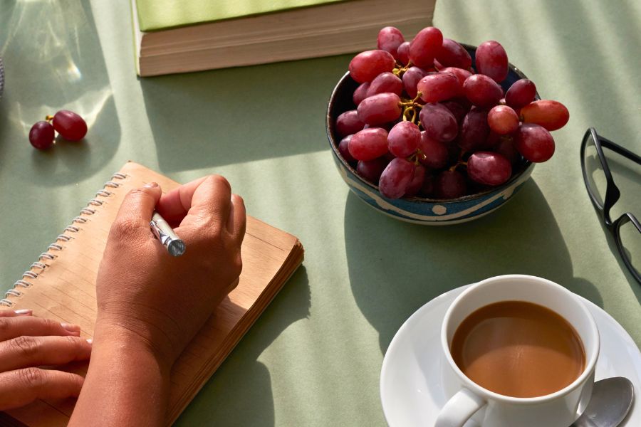 hand writing in a yellow ruled notebook, grapes in a bowl, coffee cup in a saucer
