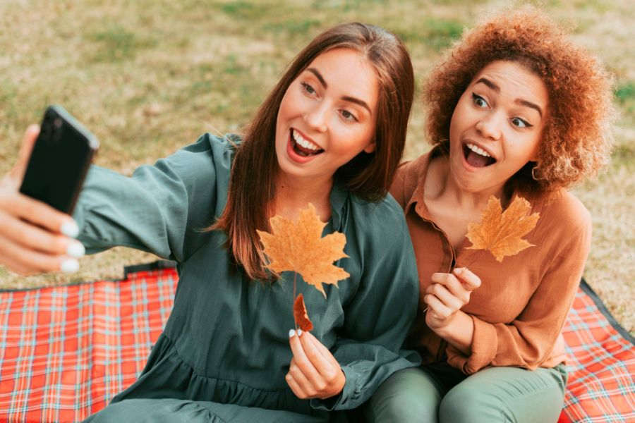 two women friends holding autumn leaf taking selfie