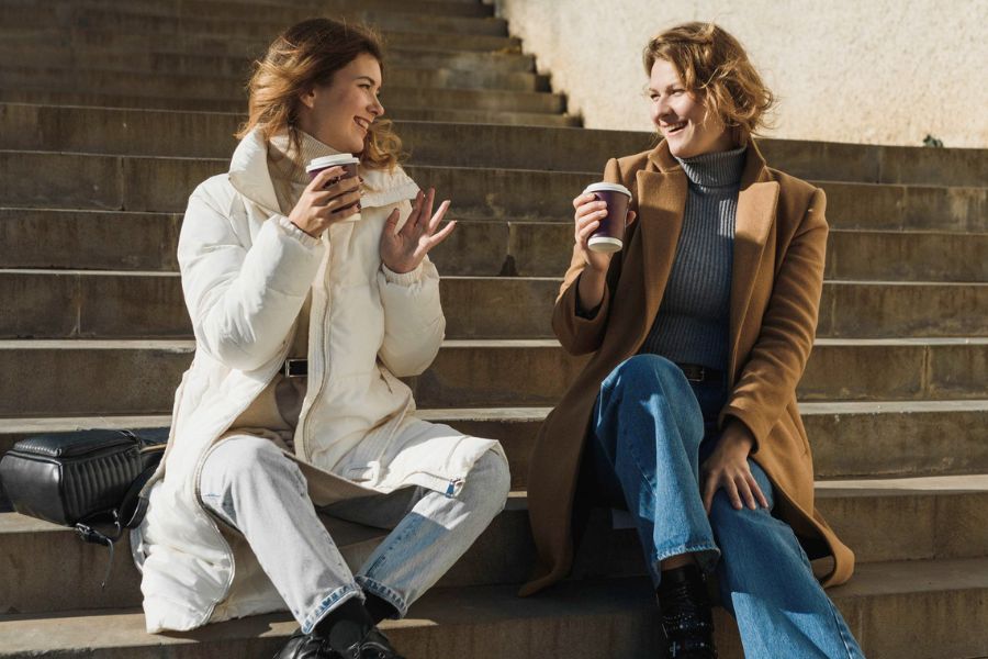 two women in winter clothes sitting on stairs talking and smiling with coffee takeout mugs in hand