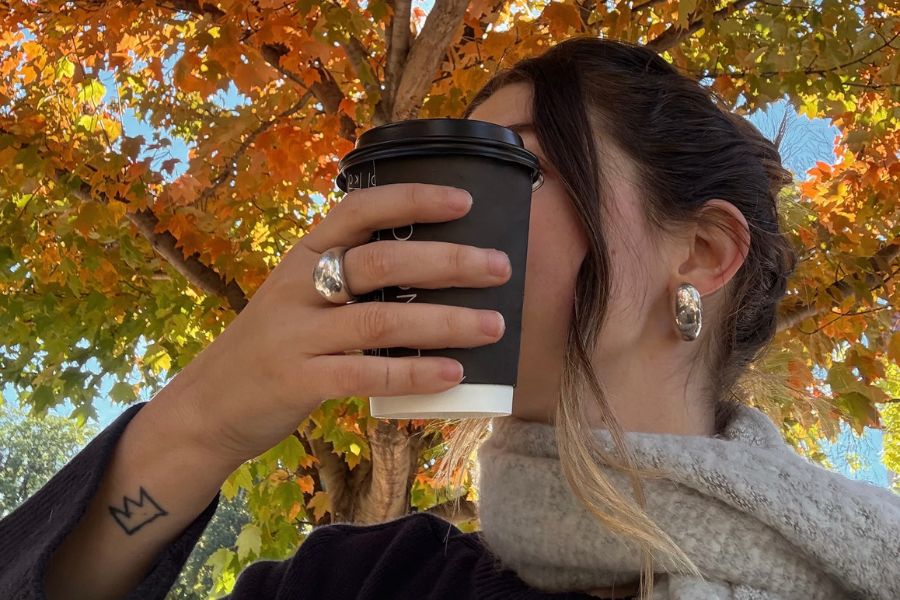 woman holding coffee takeout mug in front of her face, wearing grey scarf, with autumn tree behind her