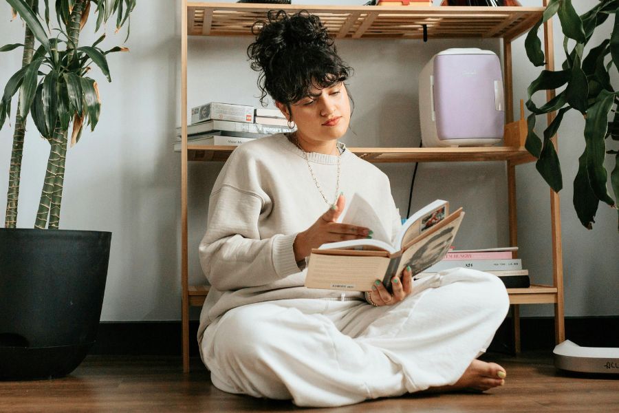 woman flipping book on floor, wearing beige sweater and white pants