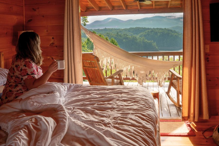 woman sitting on bed under comforter in a cottage, staring out balcony door at the view of mountains, hammock and wooden chairs in balcony