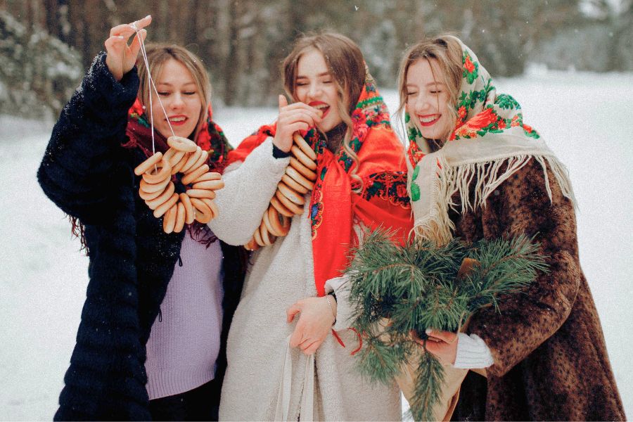 three women standing in winter clothes in snow, laughing, and eating donuts from a string